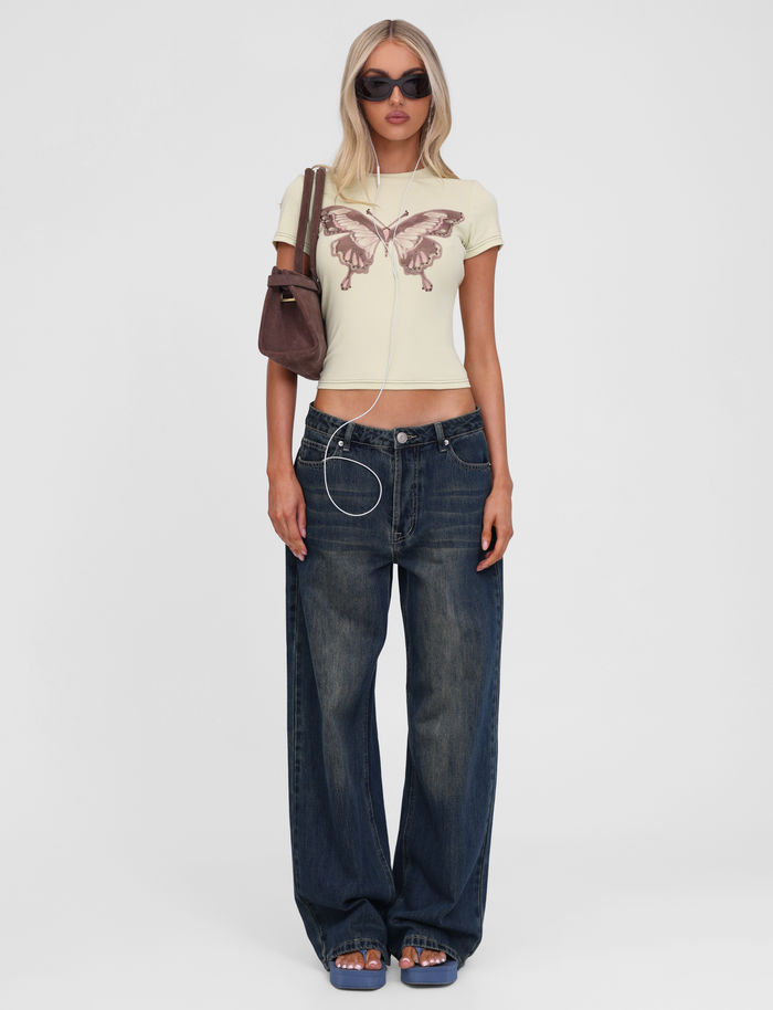 A woman wears TIGERMIST's SAPHIRA TEE in butterscotch with cap sleeves and a butterfly graphic, baggy blue jeans, blue sandals, sunglasses, a brown shoulder bag, and earphones hanging from her neck.