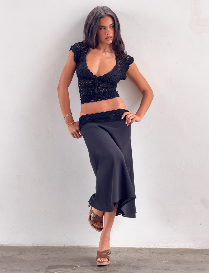 A woman with long dark hair stands against a light wall, wearing the TIGERMIST SASHA CROCHET MIDI SKIRT in black, a matching crochet top, brown heels, and gold jewelry. She looks to the side with one hand on her hip.