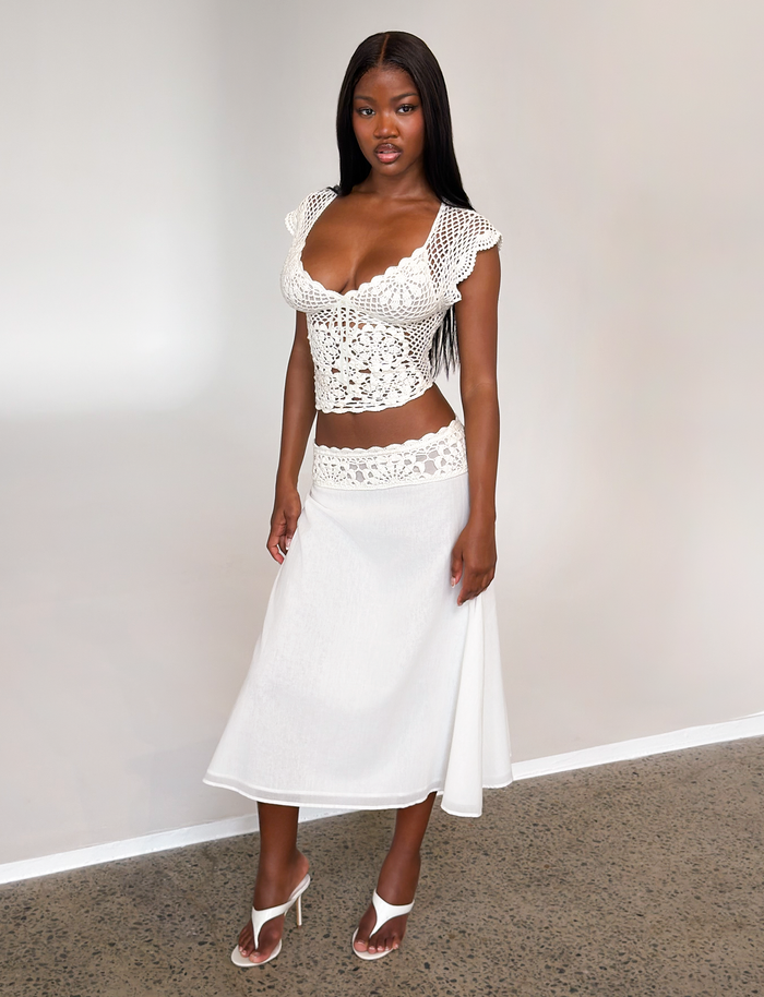 A woman stands indoors against a light background wearing the TIGERMIST SASHA CROCHET MIDI SKIRT in ivory, styled with a white crocheted cap-sleeve top and white heeled sandals.