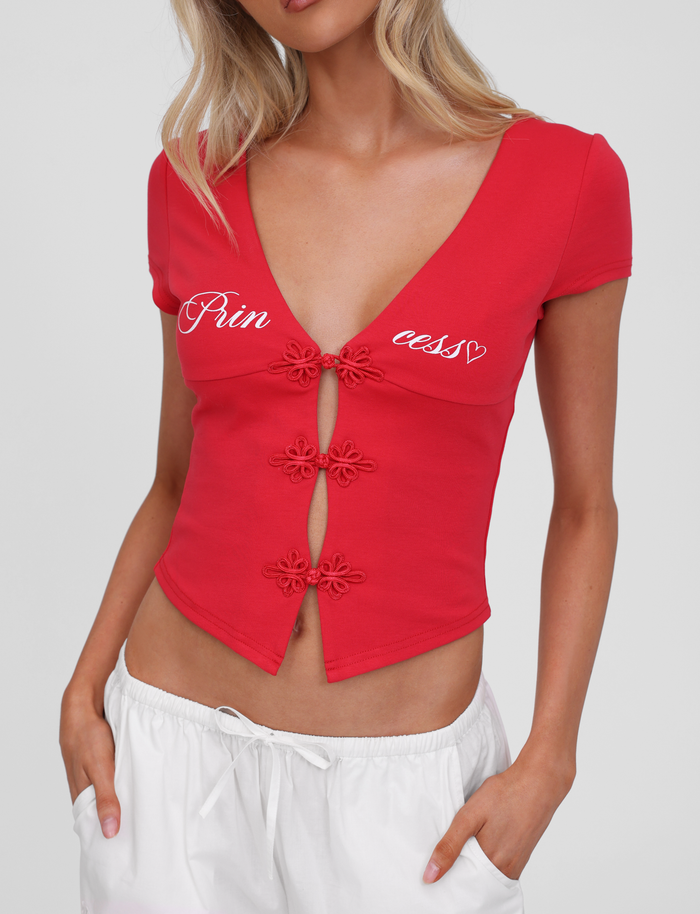 A woman wears the TIGERMIST SAVINA TOP in red, featuring embroidered buttons and a princess graphic print, paired with white pants. Her blonde hair is styled in loose waves.