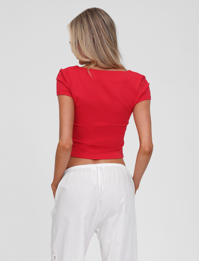 A person with long blonde hair is seen from behind against a plain light background, wearing the TIGERMIST SAVINA TOP in red with embroidered button detailing and fitted design, paired with white pants.