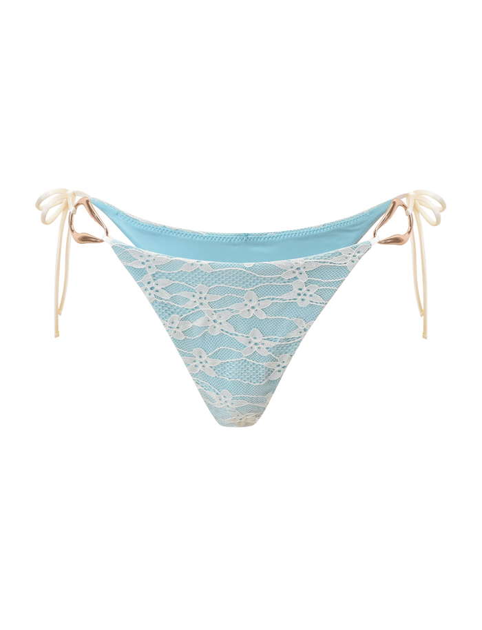 The SIKA BIKINI BOTTOM - BLUE LACE by TIGERMIST features aqua blue fabric, a white floral lace overlay, and cream side ties. With a cheeky cut, it creates a stylish summer look, shown here on a white background.