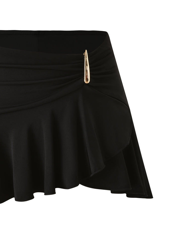 The SOLIA MINI SKIRT by TIGERMIST is a black mini skirt with a ruffled hem, gathered fabric at the waist, and a gold safety pin detail on one side.