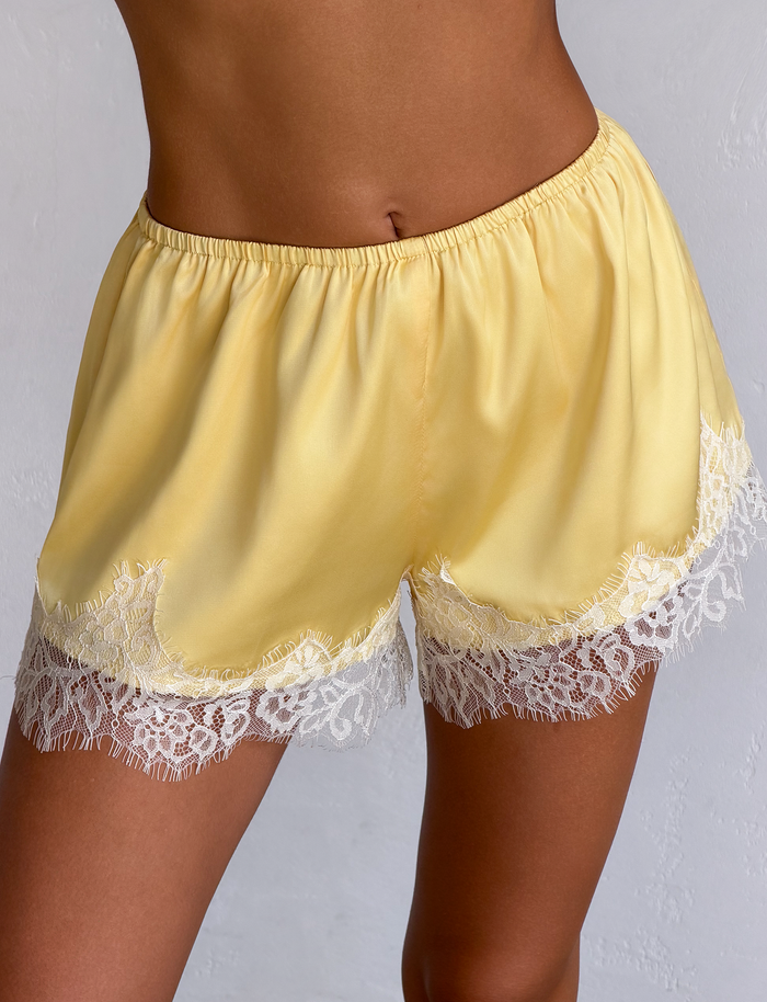 A person models the TIGERMIST SOLSTICE SHORT - YELLOW, featuring luxe satin fabric and delicate white lace trim at the hem, posed before a light-colored textured background.
