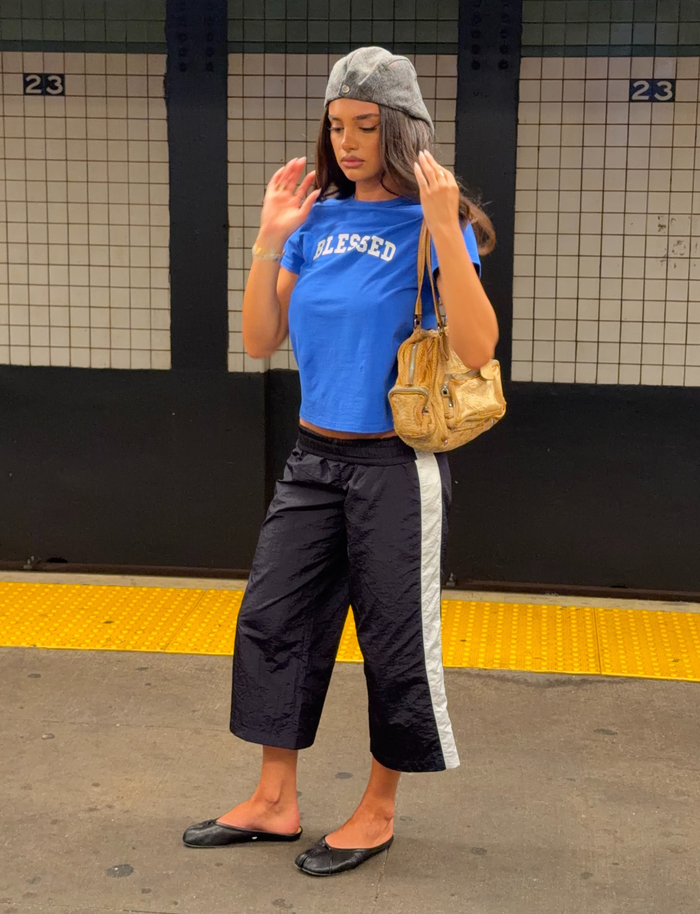 A woman stands on a subway platform wearing a gray beret, a blue “BLESSED” T-shirt, and TIGERMIST’s SOPHIA WINDBREAKER CAPRI in navy and white. She completes her look with black flats and carries a tan purse. The background features tile walls and the number 23.