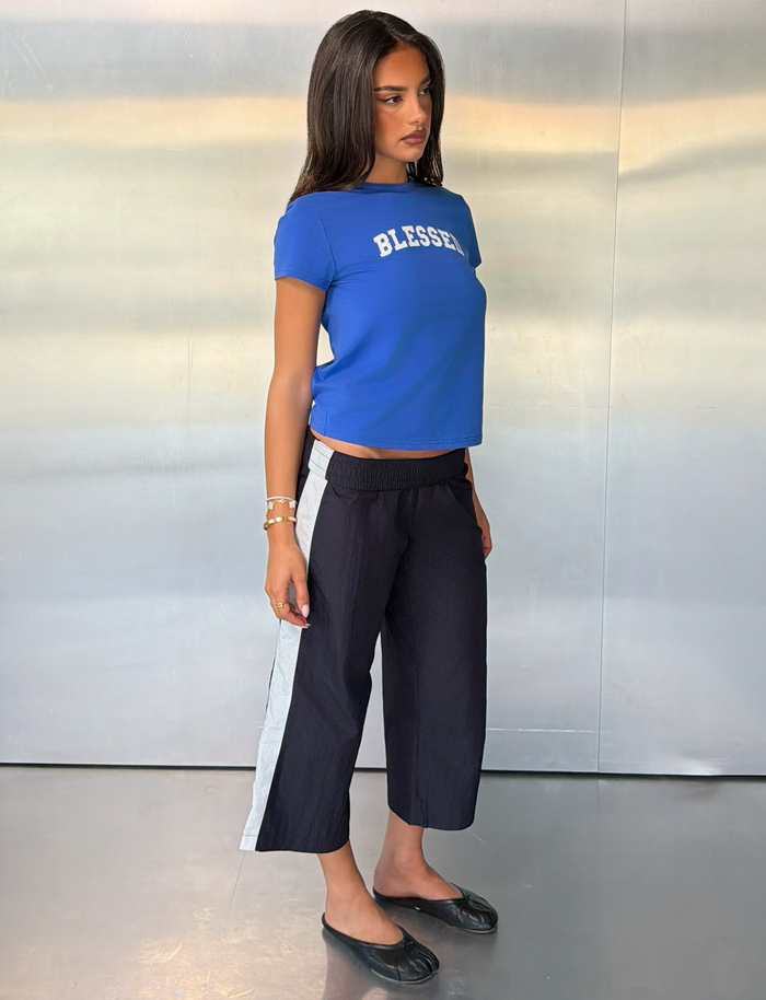 A woman stands sideways in front of a metallic wall, wearing a blue “BLESSED” T-shirt, TIGERMIST SOPHIA WINDBREAKER CAPRI pants in navy and white with wide side stripes, black flats, and bracelets on her left wrist.