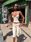 A woman wearing sunglasses, the TIGERMIST SYDEL BANDAGE TOP in cream, and matching cropped pants stands confidently on a sunny city sidewalk in front of a green storefront.