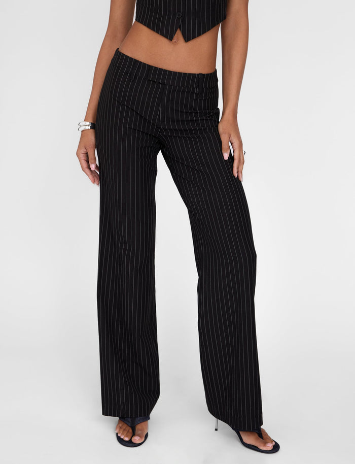 A person wearing the TAMARA PANT - BLACK : PINSTRIPE pants from TIGERMIST, featuring a mid-rise waist, paired with a matching sleeveless vest. They accessorize with bracelets adorning their wrist and black open-toed heels. The scene is set against a plain white background.