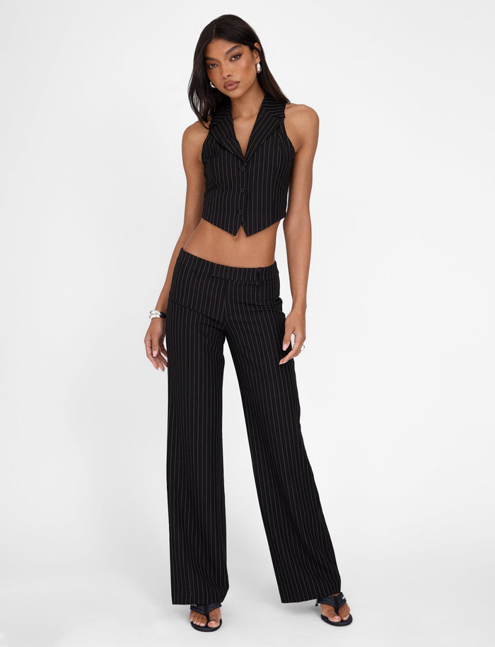 A woman strikes a confident pose in the TIGERMIST TAMARA PANT - BLACK : PINSTRIPE set, which includes a sleeveless crop vest and high-waisted, straight-leg silhouette trousers. She complements her look with bracelets, earrings, and heeled sandals while standing against a plain white background.