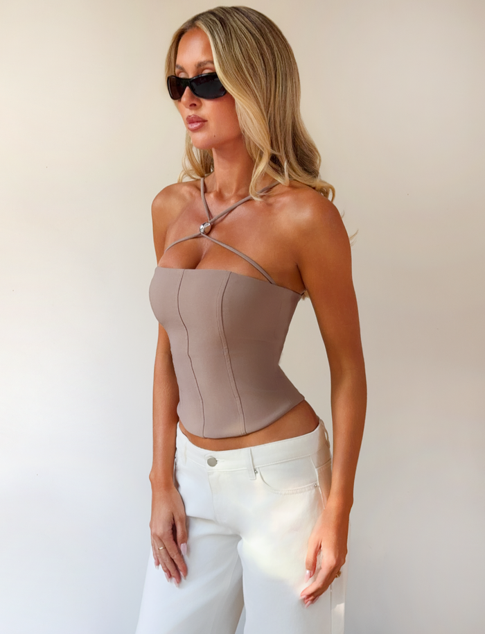 A woman with long blonde hair and black sunglasses wears the TIGERMIST TAMORA CORSET TOP in taupe with adjustable straps and white pants, standing against a plain light background.