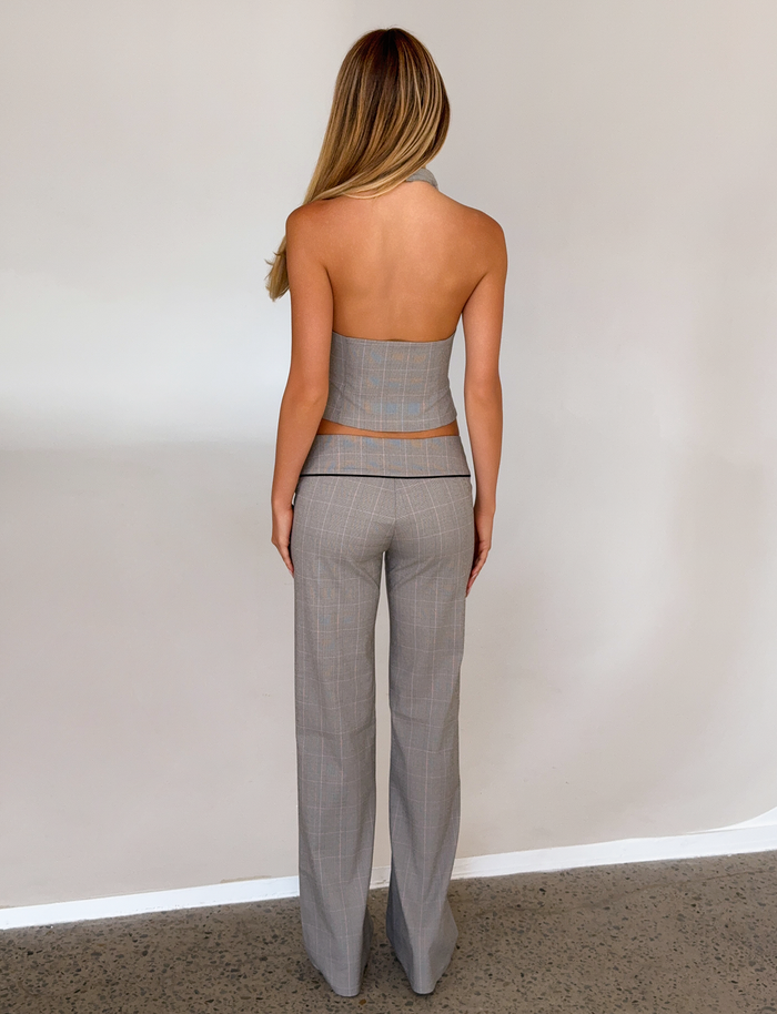 A woman with long blonde hair stands facing a light wall, wearing TIGERMIST’s TERESIA PANT in grey check with a matching strapless top. Her arms rest at her sides and her back is to the camera.