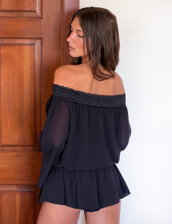 A woman with long brown hair stands by a wooden door, looking over her shoulder in the TIGERMIST TESSIE PLAYSUIT - BLACK, featuring an off-shoulder design and delicate lace trim.