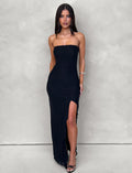 A woman with long dark hair is standing against a light gray background, wearing the black THEO MAXI DRESS by TIGERMIST, which features a thigh-high slit on one side. She pairs it with black heeled sandals. Her hands are by her sides, and she has a neutral expression on her face.