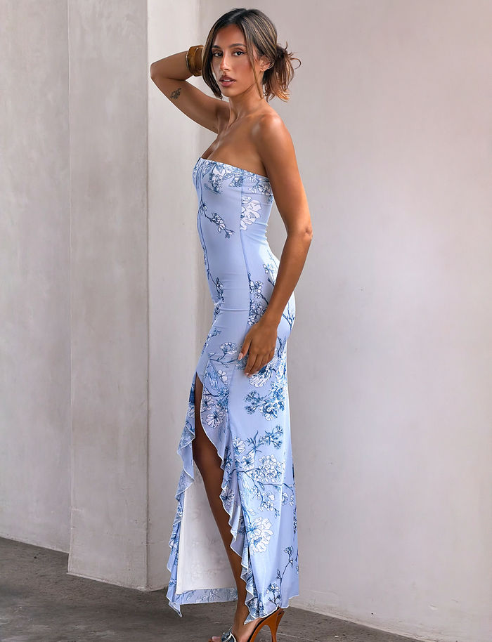 A woman stands sideways against a light wall, looking at the camera, wearing TIGERMIST’s THEO MAXI DRESS in blue floral mesh with a thigh-high split and ruffle detail, paired with high-heeled sandals.