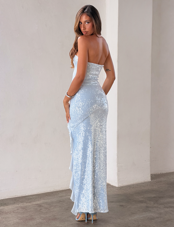 A woman in the TIGERMIST THEO SEQUIN MAXI DRESS - SOFT BLUE, featuring a mermaid silhouette, poses on a concrete floor with long hair and high heels in a minimalist, light-toned setting, turning her head toward the camera.