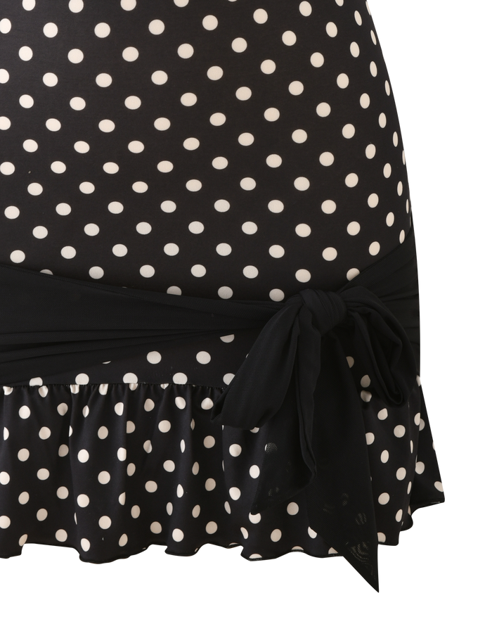 A close-up of the TOVE MINI DRESS - BLACK POLKA DOT by TIGERMIST, featuring white polka dots, a ruffle hem, and a black tie at the waist for a playful touch on the bodycon silhouette.