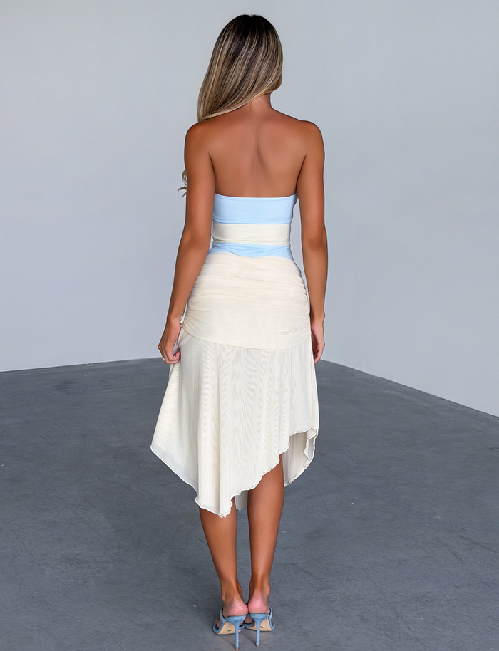 A woman with long blonde hair stands indoors, facing away, wearing the TIGERMIST TWYLA MIDI DRESS in cream and blue, paired with blue high-heeled sandals on a gray floor against a plain light-colored wall.