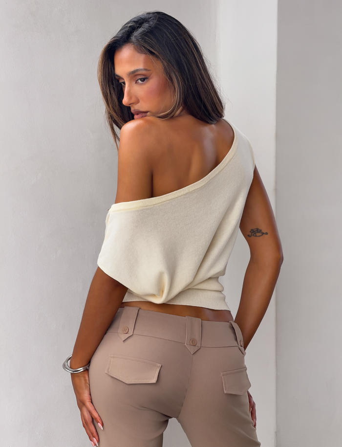A woman with loose hair and a bracelet looks over her shoulder, wearing the TIGERMIST TYLA SLEEVELESS KNIT TOP in cream with beige pants; she has a small upper arm tattoo.