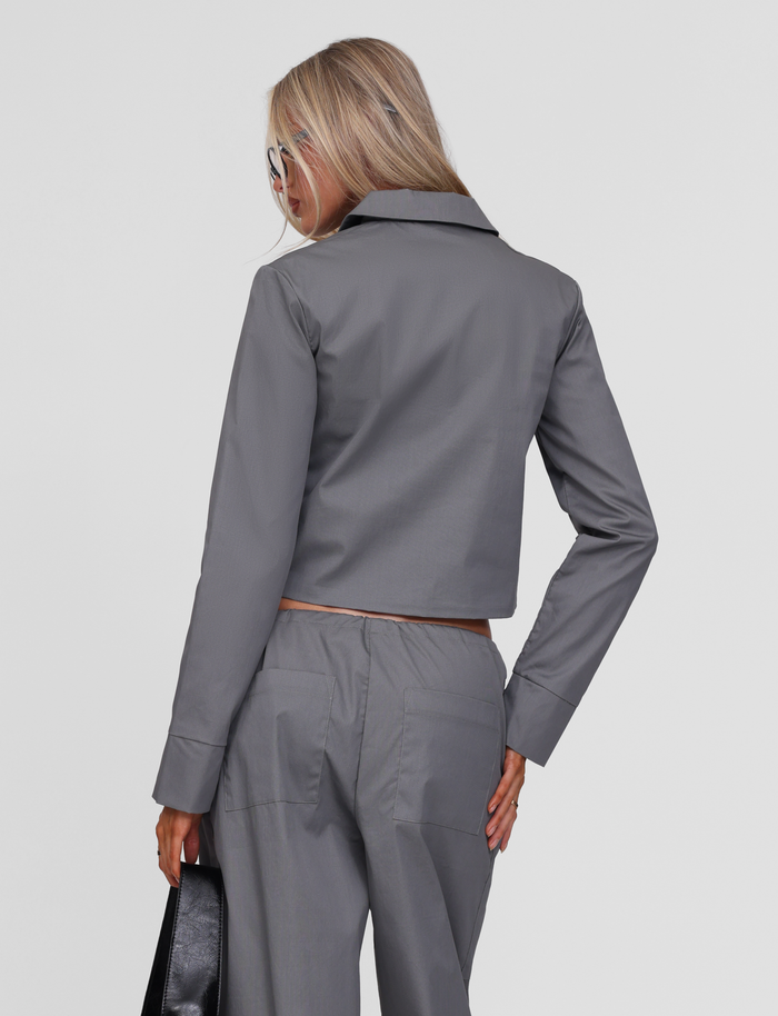 A woman with long blonde hair and sunglasses wears the TIGERMIST TYSHA JACKET in charcoal with matching pants, facing away from the camera and holding a black handbag in her left hand.