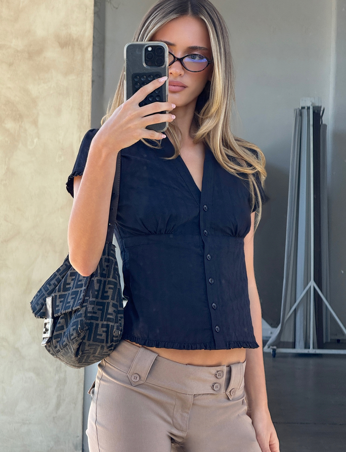 A woman with long, light brown hair wears cat-eye sunglasses, beige pants, and the TIGERMIST VALERIE SHORT SLEEVE SHIRT in black. She holds a phone and has a patterned shoulder bag while taking an indoor mirror selfie.