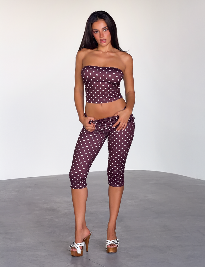 A woman with long, dark hair poses confidently in a minimalistic room, wearing the TIGERMIST VALI POLKA DOT CAPRI - CHOCOLATE PINK DOT and a strapless maroon top, paired with white high-heeled sandals.