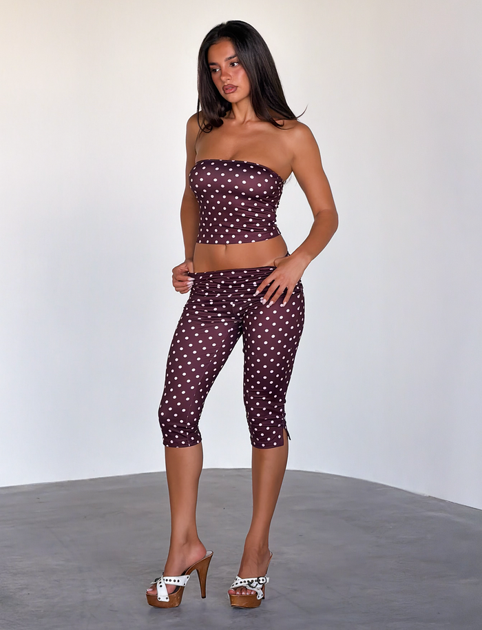 A woman stands on a gray floor wearing the TIGERMIST VALI POLKA DOT CAPRI in Chocolate Pink Dot with a strapless brown crop top and white high-heeled sandals. The background is light and plain.