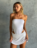 A woman with long blonde hair wears the CL - VERITY SKORT by Tiger Mist USA and a strapless white top, standing against a gray textured background. She looks slightly down and to the side with a relaxed expression.