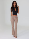 A woman with long hair stands against a neutral background, showcasing a stylish full-length silhouette. She is wearing a sleeveless black top with a keyhole cutout and the VESPER PANT in tan from TIGERMIST, featuring a low-rise waist. Black heels complete her look as one hand rests on her hair while the other hangs by her side.