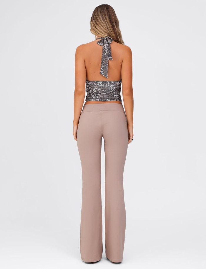 A woman with long hair stands with her back to the camera, showcasing a full-length silhouette in a halter top with leopard print and TIGERMIST's VESPER PANT - BROWN : TAN. The background is plain and white.