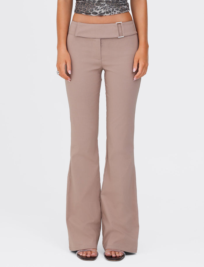 A person stands against a white background wearing the VESPER PANT - BROWN : TAN by TIGERMIST. The taupe-colored flare pants feature a wide waistband and a rectangular belt buckle. The solid tan pants have a full-length silhouette that complements their patterned cropped top. They are wearing sandals, with their hands resting by their sides.