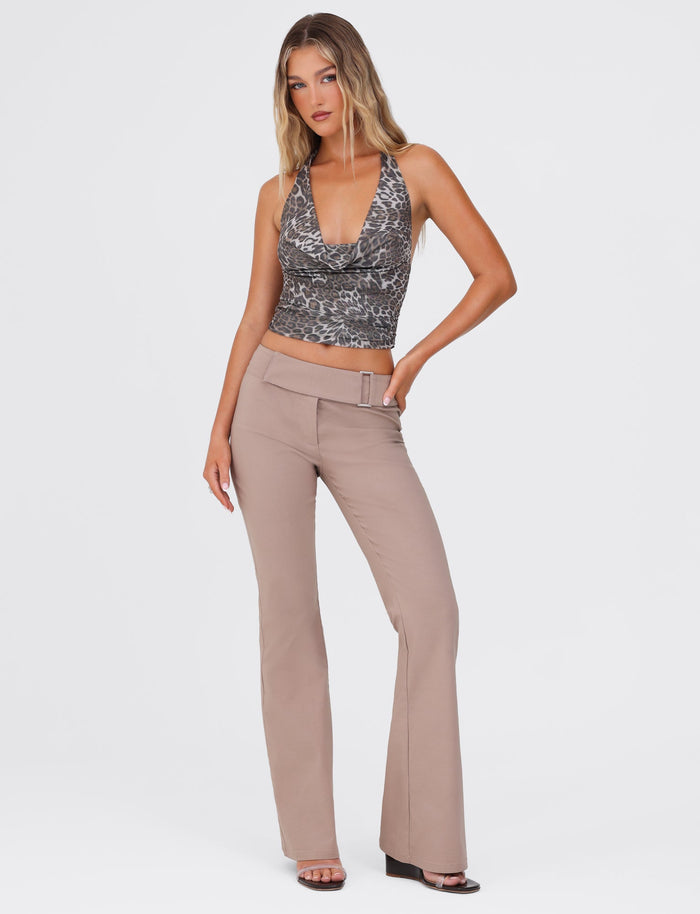 A woman with long blonde hair stands against a plain white background. She is wearing a sleeveless, low-cut leopard print top and TIGERMIST's VESPER PANT in brown tan with a belt detail. She is posing with one hand on her hip and wearing black heels.