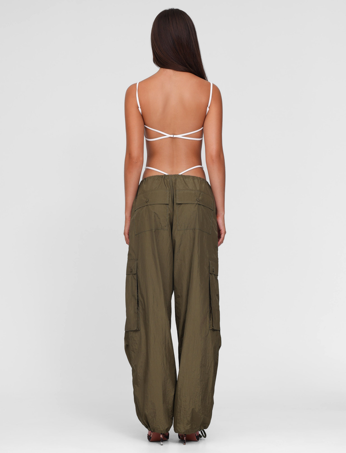 A woman faces away, wearing a strappy white open-back top and TIGERMIST VIOLETTE PANT in khaki green—loose, low-rise cargo pants with multiple pockets—against a plain light gray background.