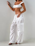 A woman poses against a white wall wearing TIGERMIST's VIOLETTE PANT in white, styled with a cropped off-shoulder top and black open-toe heels. Her face is not visible.