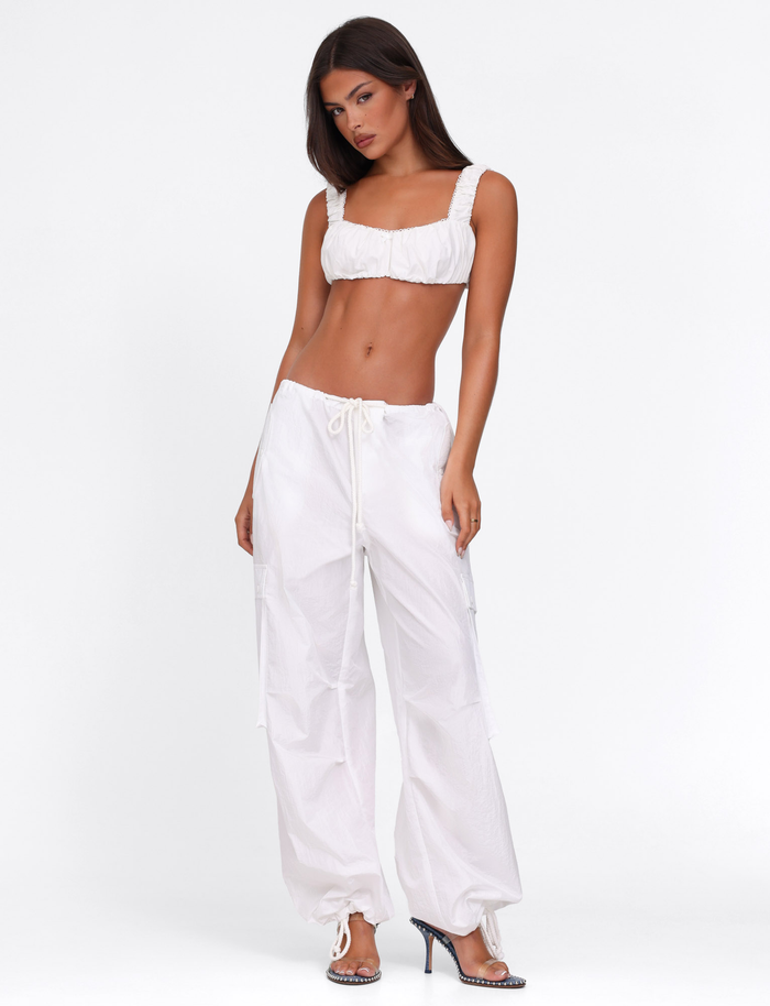 A woman poses against a white background wearing the TIGERMIST VIOLETTE PANT in white, paired with a white crop top and high-heeled shoes. She stands slightly angled, looking confidently at the camera.