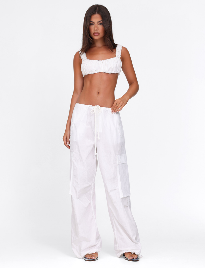 A woman with straight brown hair and a neutral expression wears the TIGERMIST VIOLETTE PANT - WHITE, an oversized fit, low rise nylon cargo pant with drawstrings, styled with a white crop top and black sandals against a plain white background.