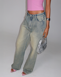 A person wears a pink sleeveless top, ZANTHE JEAN - GREY WASH by TIGERMIST, pink open-toe heels, a silver bracelet, and holds a metallic silver handbag against a plain light background.