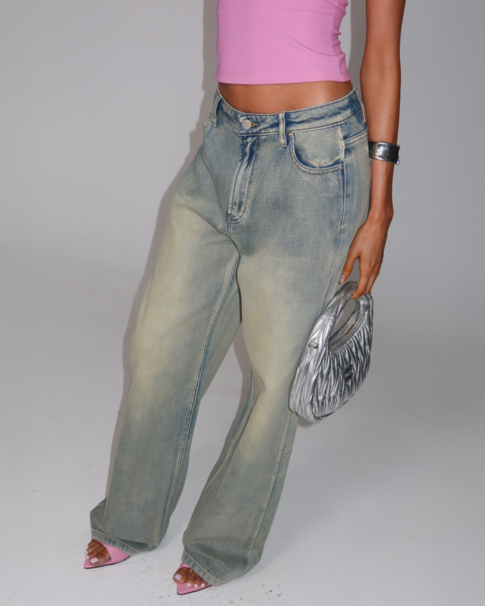 A person wears a pink sleeveless top, ZANTHE JEAN - GREY WASH by TIGERMIST, pink open-toe heels, a silver bracelet, and holds a metallic silver handbag against a plain light background.