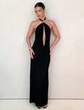 A woman stands against a light wall, wearing the TIGERMIST ZARA MAXI DRESS in black. The dress features a halter neckline, front keyhole cutout, and elegant open back. She styles her hair back and accessorizes with earrings and a bracelet.