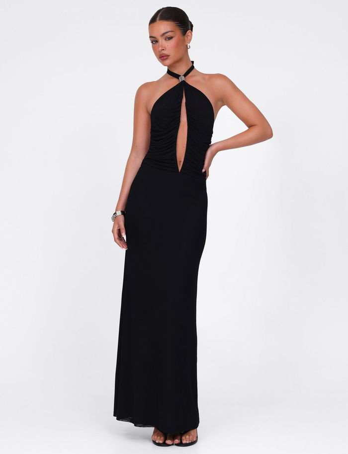 A woman models the TIGERMIST ZARA MAXI DRESS - BLACK, featuring a sleek halter neckline and front keyhole cutout, styled with black heels and bracelets against a plain white background. Her hair is tied back.