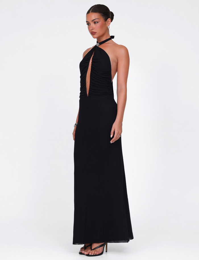 A woman stands on a white background wearing the TIGERMIST ZARA MAXI DRESS in black, featuring a long, sleeveless halter design with a deep front cutout. Her hair is in a neat bun and she pairs the dress with black high-heeled sandals.