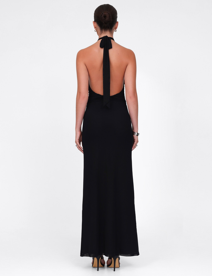 A woman with a neat bun and a bracelet on her left wrist wears the TIGERMIST ZARA MAXI DRESS in black, featuring a halter neck, open back, and neck tie. She pairs it with tan heels, standing against a plain white background.
