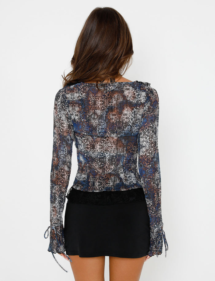 A person with long brown hair is facing away from the camera, styled in the semi-sheer, long-sleeved TIGERMIST ZELDA TOP in black abstract shades of blue, gray, and white. The blouse features ruffled shoulder details and pairs beautifully with their black skirt. The background remains a plain white.