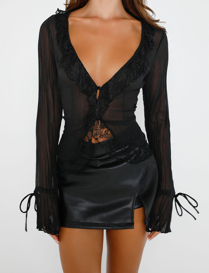 A woman is wearing the ZELDA TOP - BLACK from TIGERMIST, a sheer black long-sleeve top with lace detailing and a deep V-neckline, paired with a black leather mini skirt featuring a small slit on the left side. She stands against a plain white background.