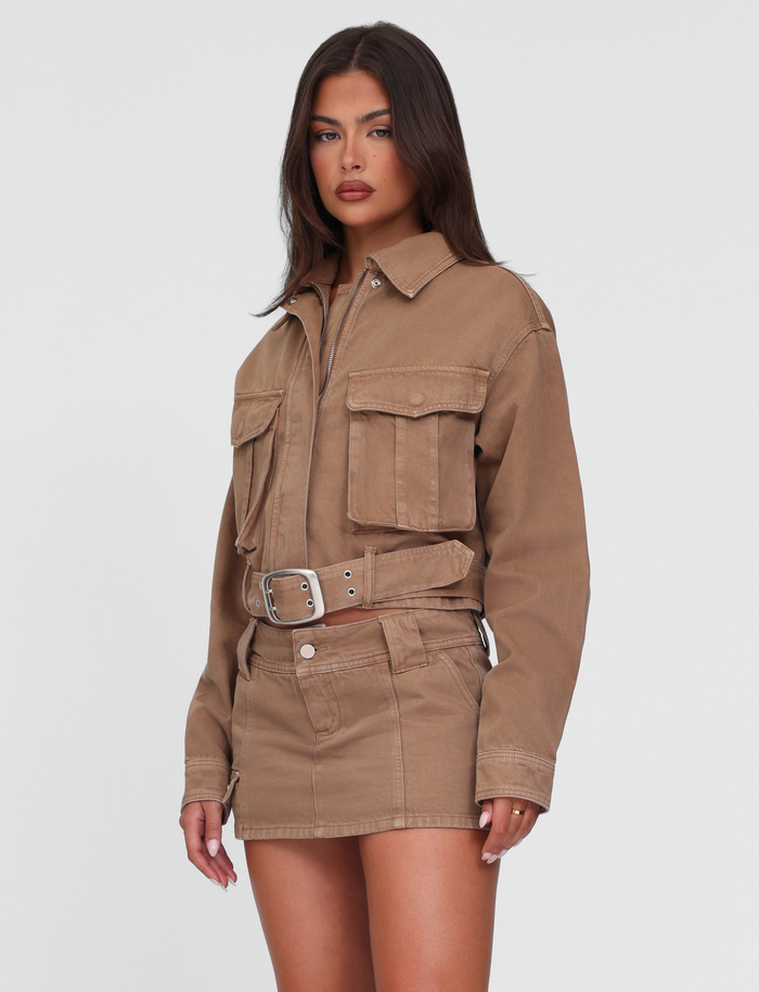 A woman with long dark hair and a neutral expression wears the TIGERMIST ZELIE JACKET in Sandblast Washed Tan, styled with a matching mini skirt, standing against a plain white background.