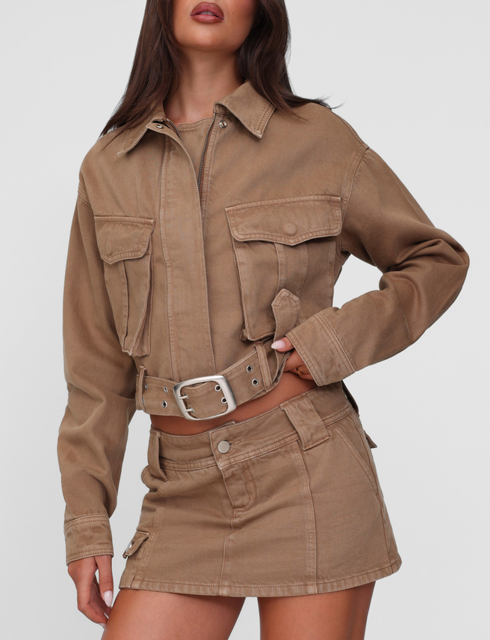 A woman wears the TIGERMIST ZELIE MINI SKORT in washed tan, styled with a tan cropped jacket featuring large front pockets and silver buttons. The skort has seam details, functional hip pockets, and an asymmetrical design.
