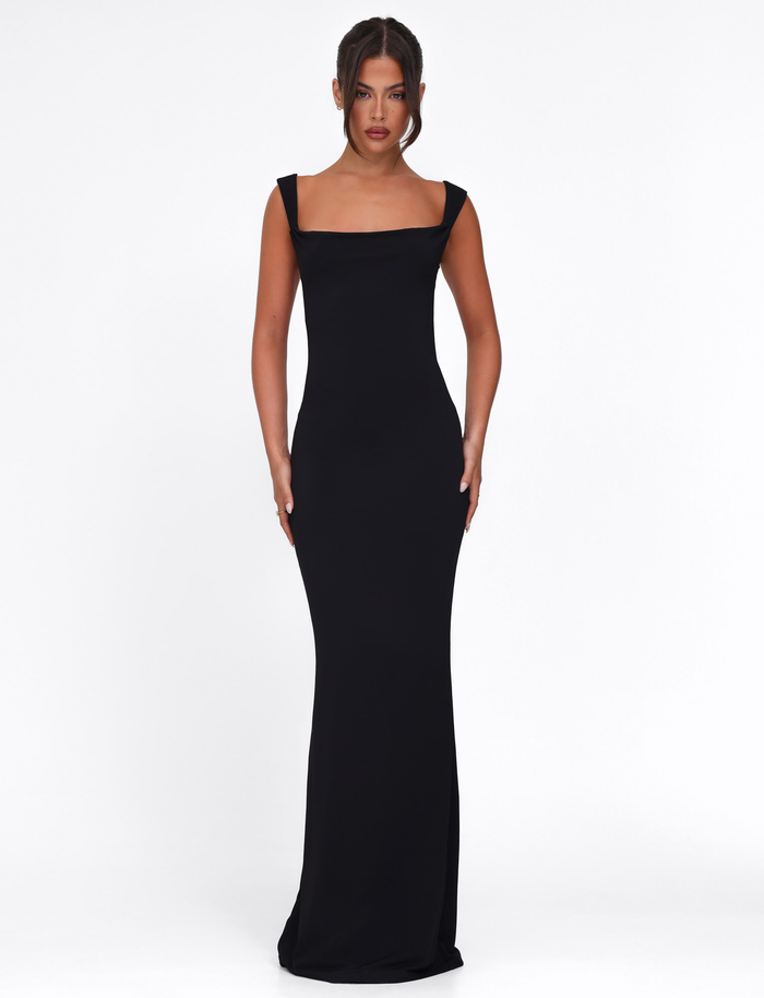 A woman faces forward in the TIGERMIST ZITA DRESS - BLACK, a bodycon maxi with wide shoulder straps. Her hair is in a neat updo as she poses against a plain white background.