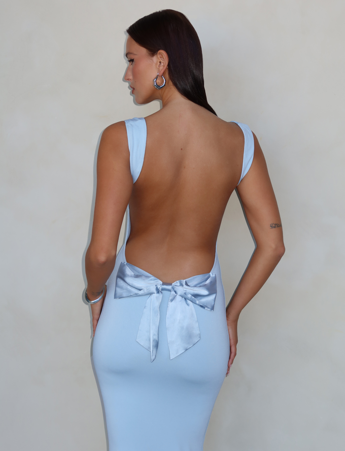 A woman with straight dark hair wears the TIGERMIST ZITA DRESS - BLUE: BABY BLUE, a backless bodycon style featuring a large satin bow at the lower back, as she stands facing away against a plain, neutral background.