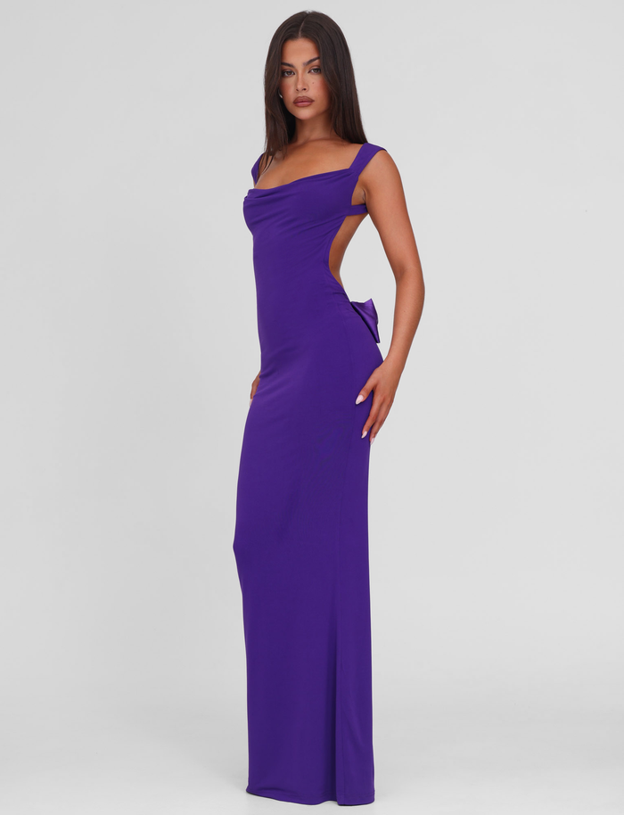 A woman with long dark hair models the TIGERMIST ZITA MAXI DRESS in purple, featuring a sleeveless bodycon silhouette, open back, and side cutout, standing against a plain white background.