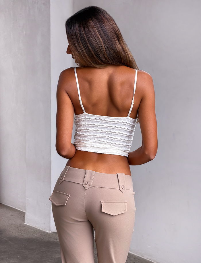 A woman with long brown hair, wearing the TIGERMIST ZORA TOP in cream with adjustable straps and beige pants, stands with her back to the camera against a light-colored wall.